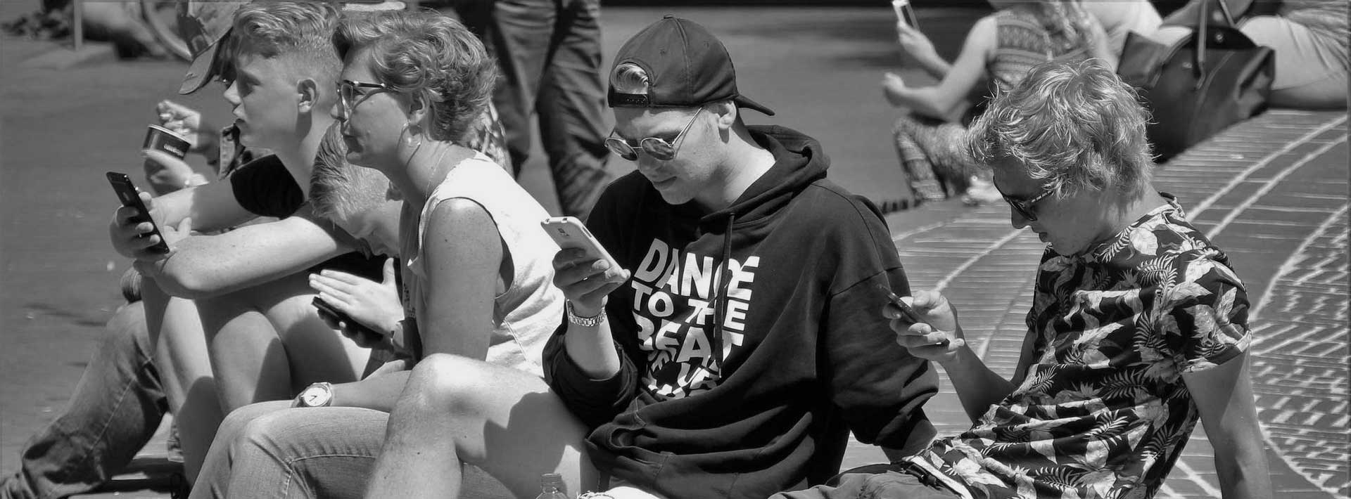 Obraz przedstawia młodzież siedzącą na chodniku. Każdy z młodzieńców wpatrzony jest w telefon komórkowy